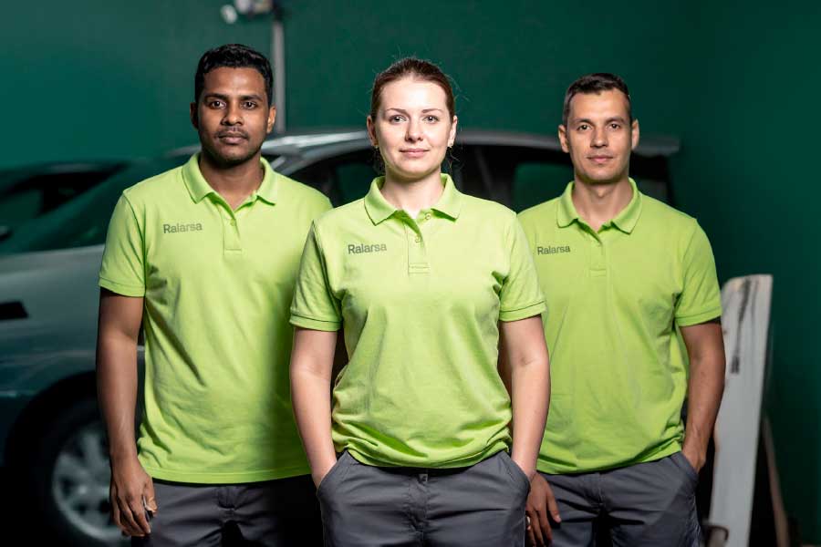 Tres técnicos uniformados con ropa corporativa de Ralarsa, de pie frente a un taller de autos, simbolizando trabajo en equipo y solidez empresarial.